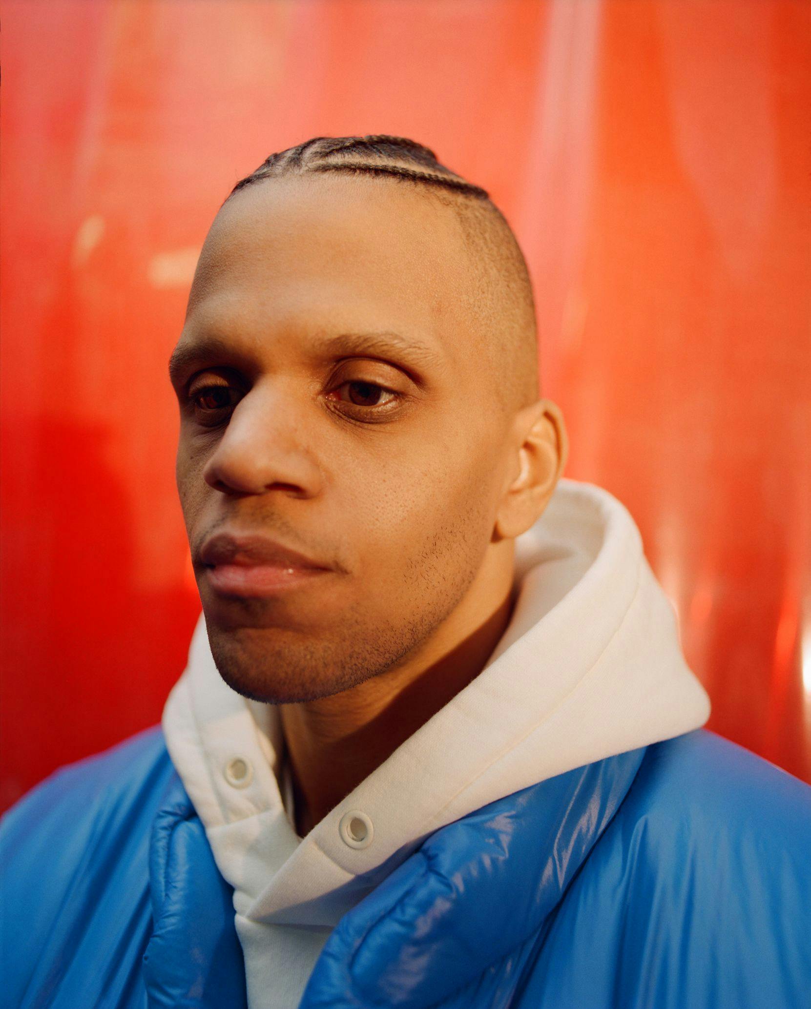 A close-up view of a brown-skinned person wearing a white hoodie and blue jacket in front of a red welding screen.