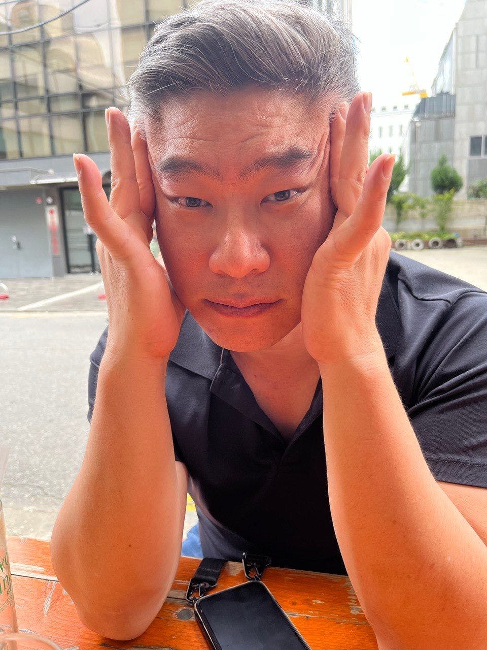 A man seated at a table against a backdrop of a city square holds his face between his hands and looks into the camera.