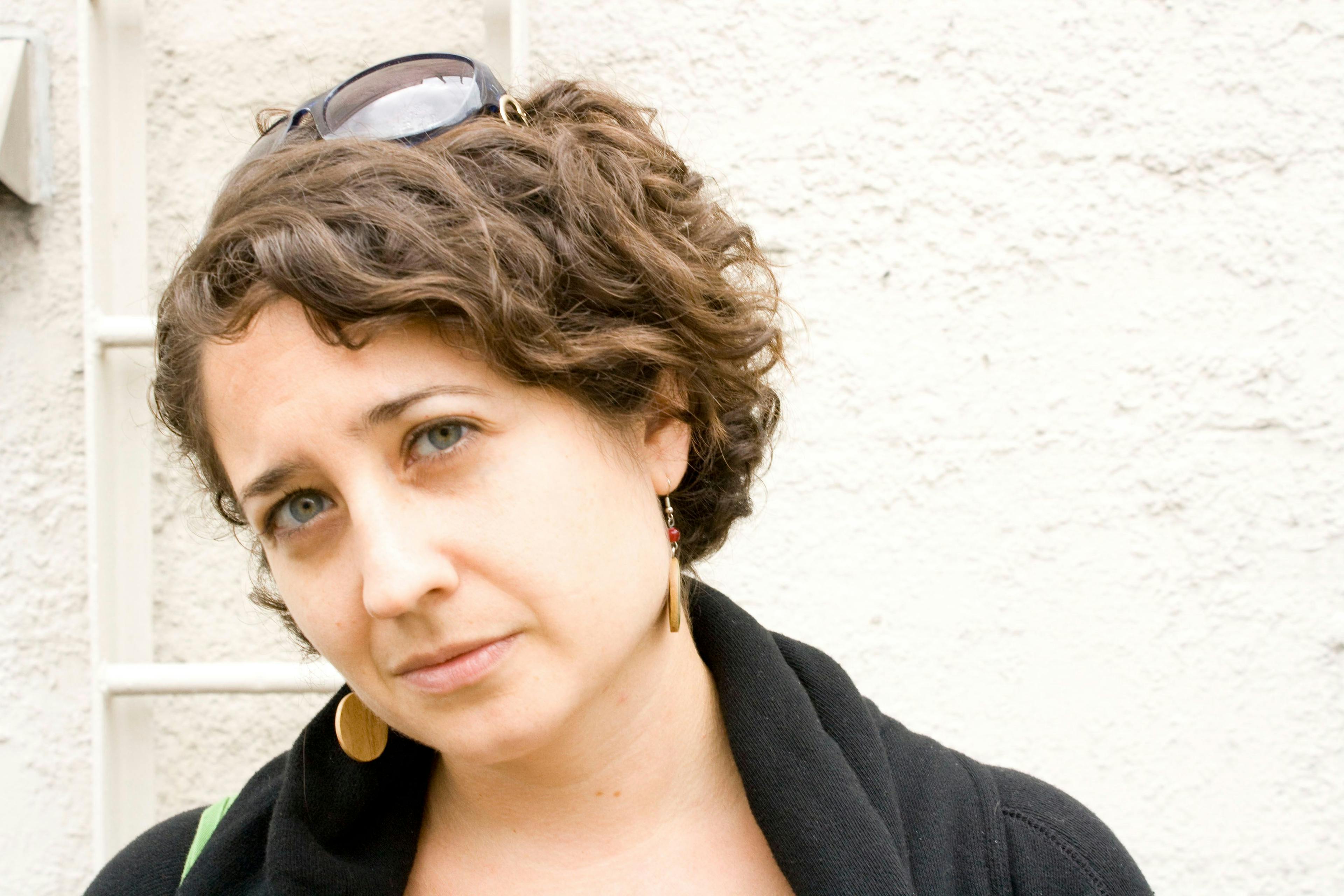 A white woman with wavy brown hair and hazel eyes looks up towards the camera, her head slightly tilted. She wears a black top, dangly wooden earrings, and sunglasses on her head.